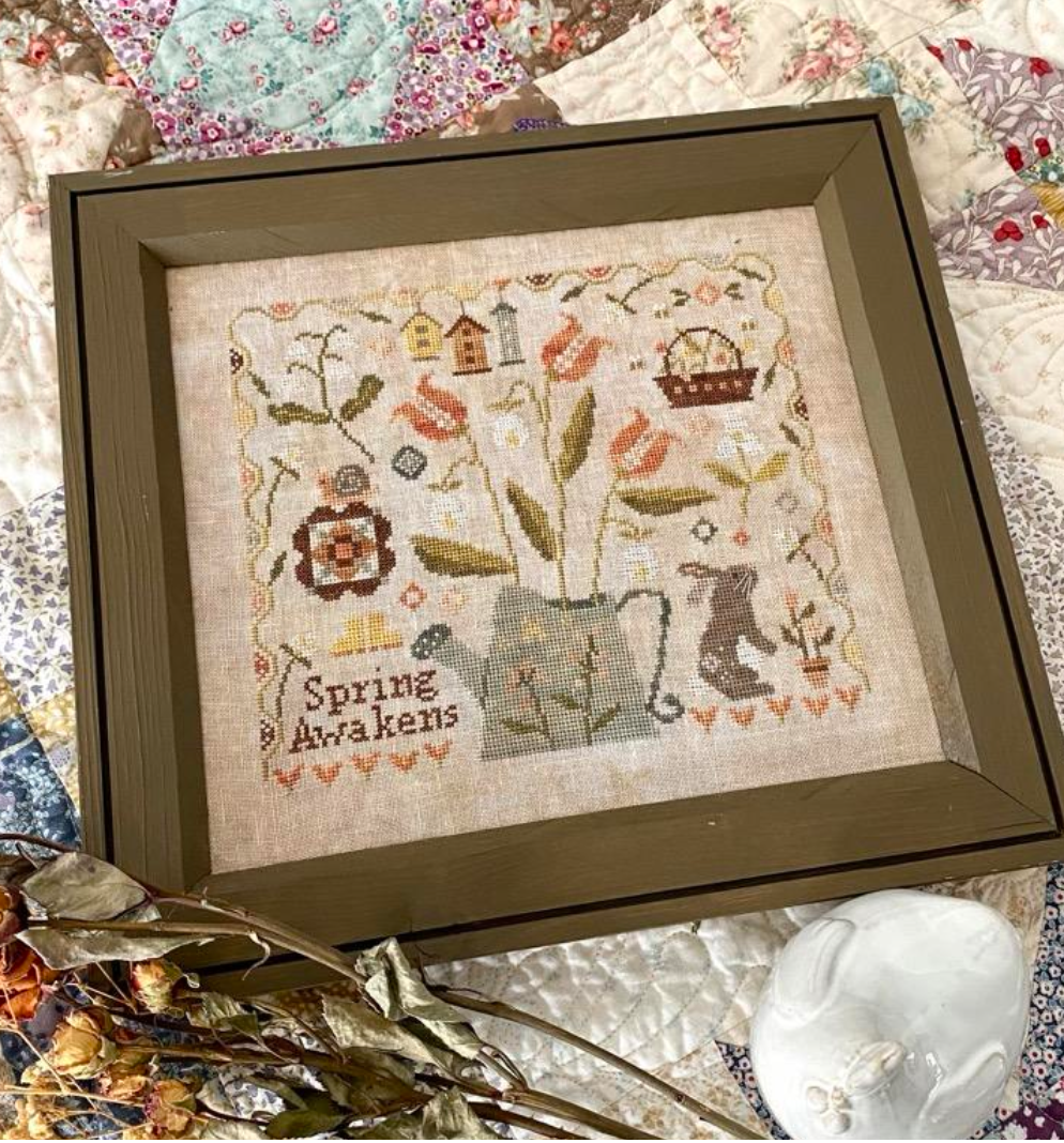 Framed cross-stitch art with 'Spring Awakens' text on a textured fabric background