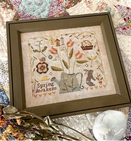 Framed cross-stitch art with 'Spring Awakens' text on a textured fabric background