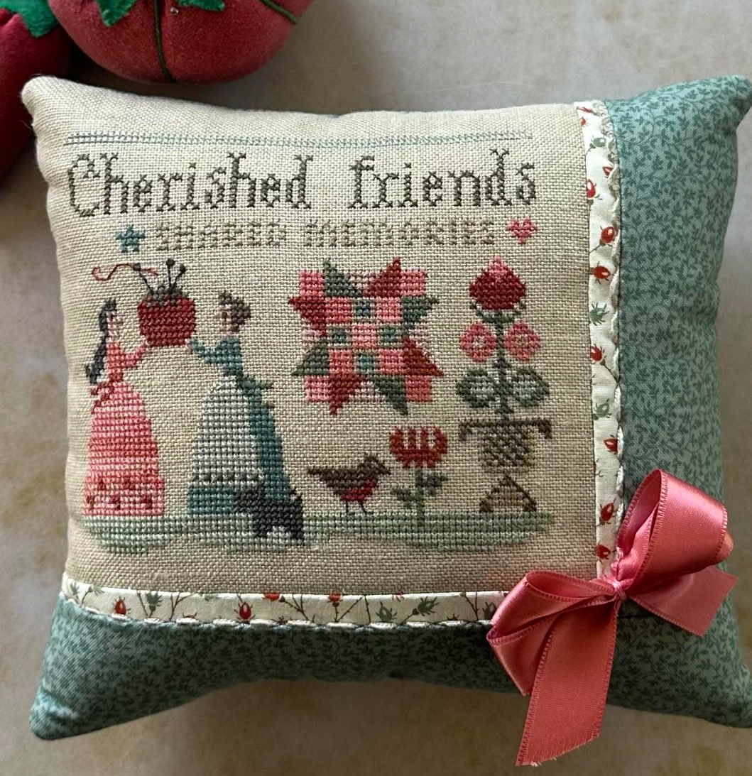 Decorative pillow with cross-stitch design and text 'Cherished friends share memories' on a wooden surface.